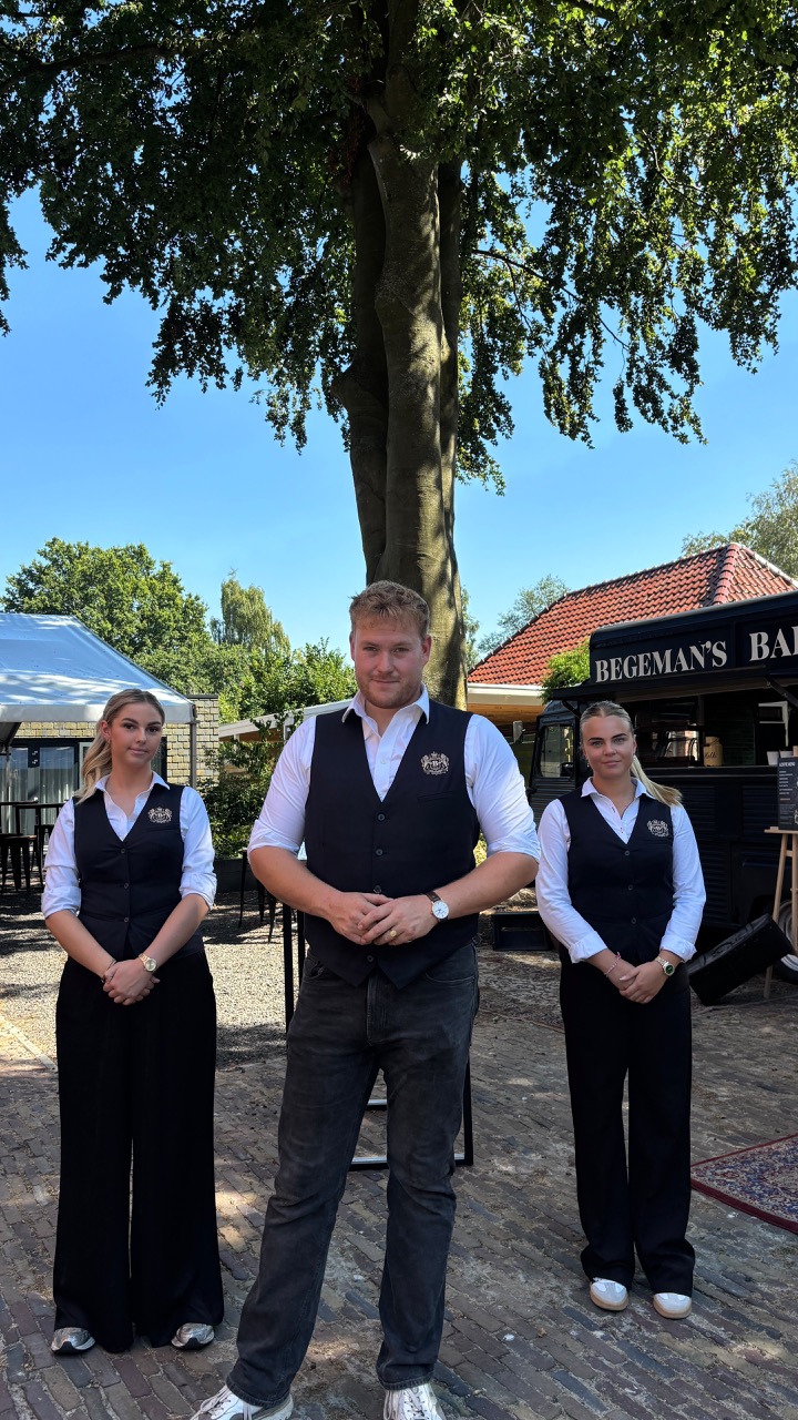 Drie horecamedewerkers poseren buiten onder een grote boom
